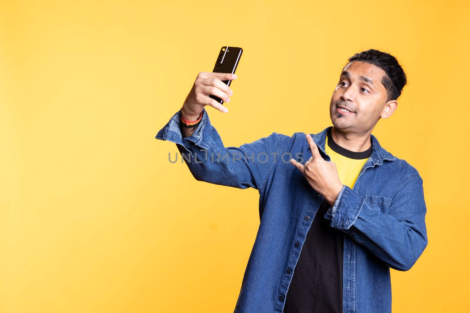 Young adult takes photos on his smartphone while he wears colorful clothes, showing a peace sign in front of the camera. Smiling indian person with man bum using his mobile phone in studio.