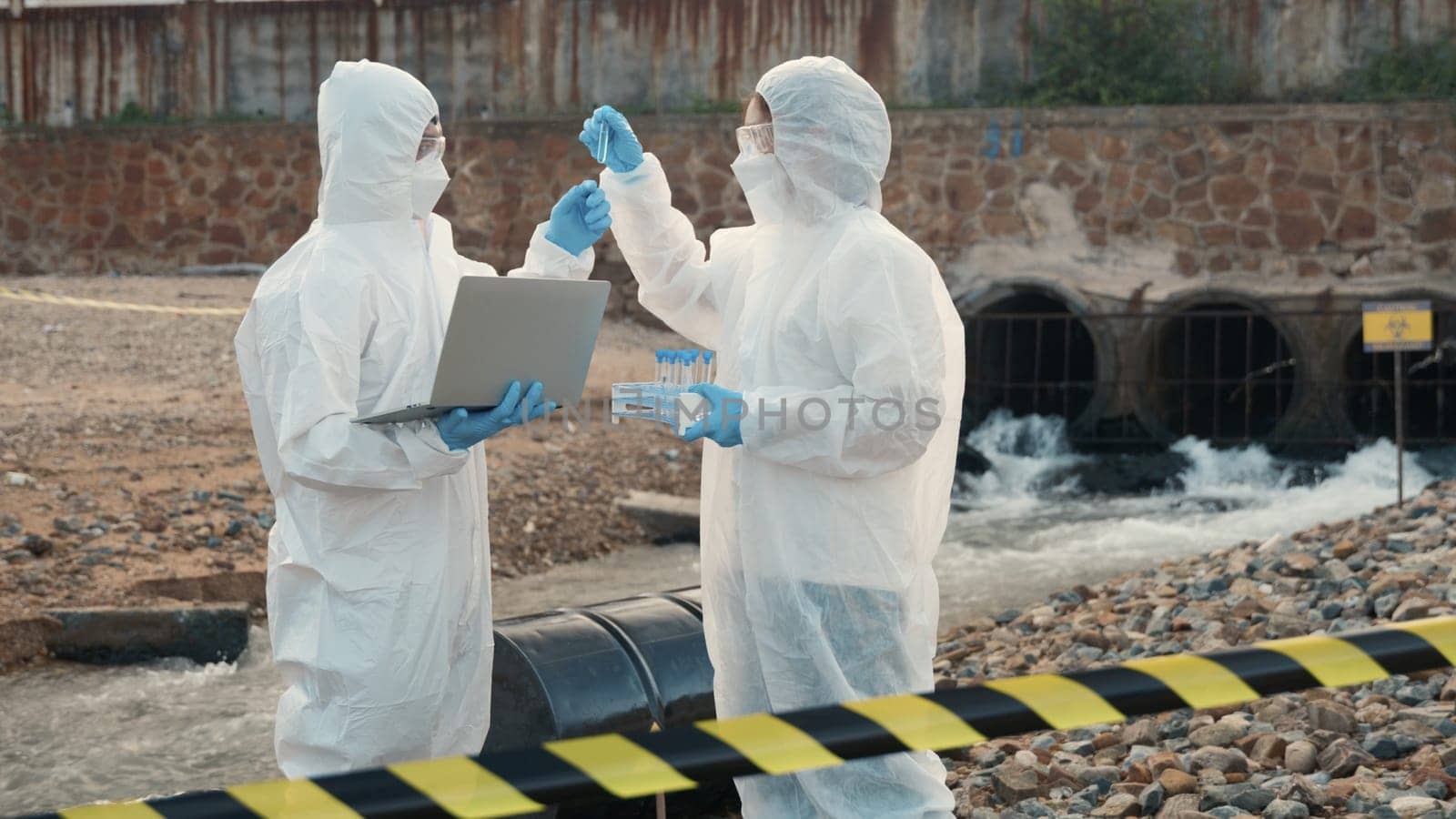 Ecologist sampling water from the river with test tube and save data to laptop computer, Biologist in a protective suit and mask collects sample of waste water from industrial, problem environment