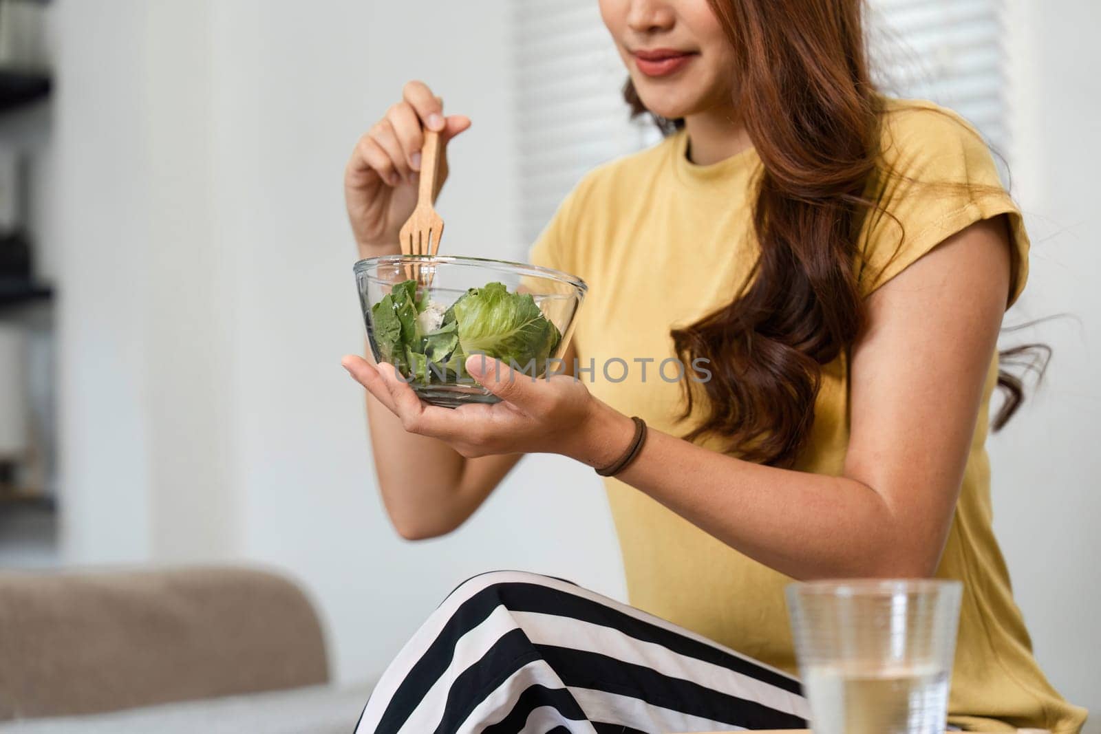 In her cozy living room, a woman takes pleasure in a fresh salad, embracing her healthy morning routine and nurturing her body with wholesome food.