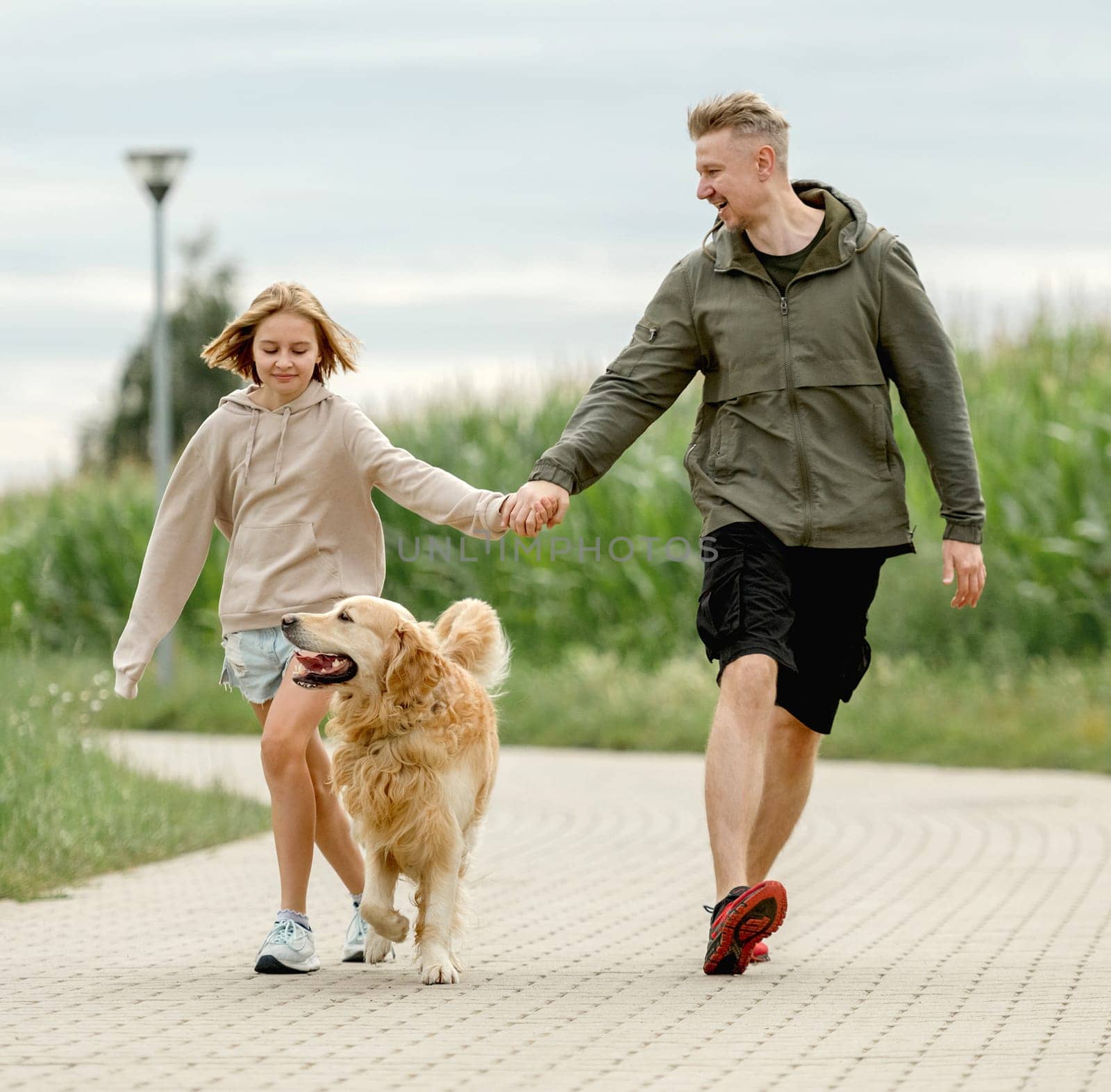 Dad With Daughter And Golden Retriever Dog Run Along The Path