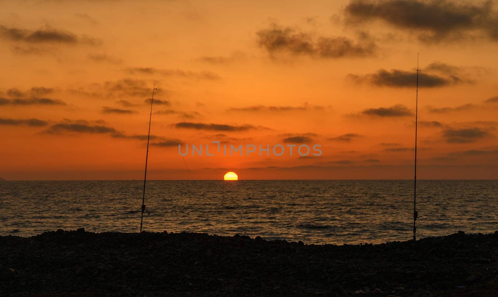Two fishing rods stand tall against the vibrant sunset, as waves gently lap at the rocky shoreline, creating a peaceful atmosphere perfect for evening leisure.