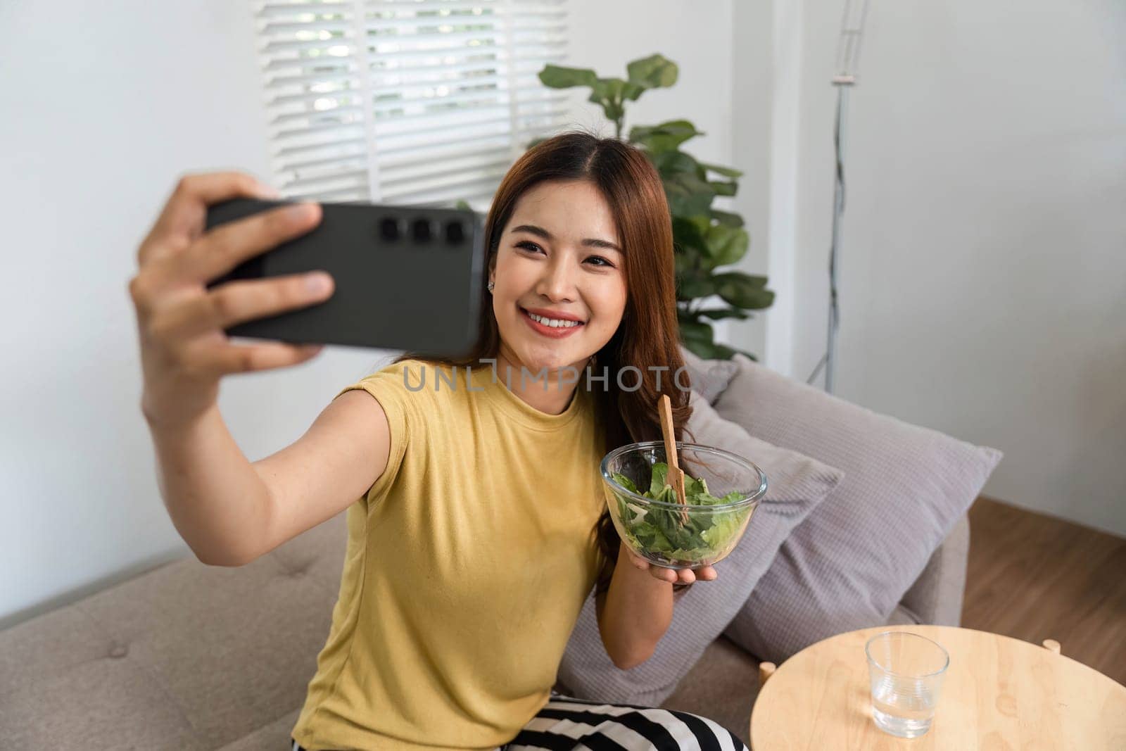 A cheerful woman captures her morning routine by taking a selfie with a healthy bowl of salad. This moment showcases her dedication to wellness and self-care in a comfortable home environment.