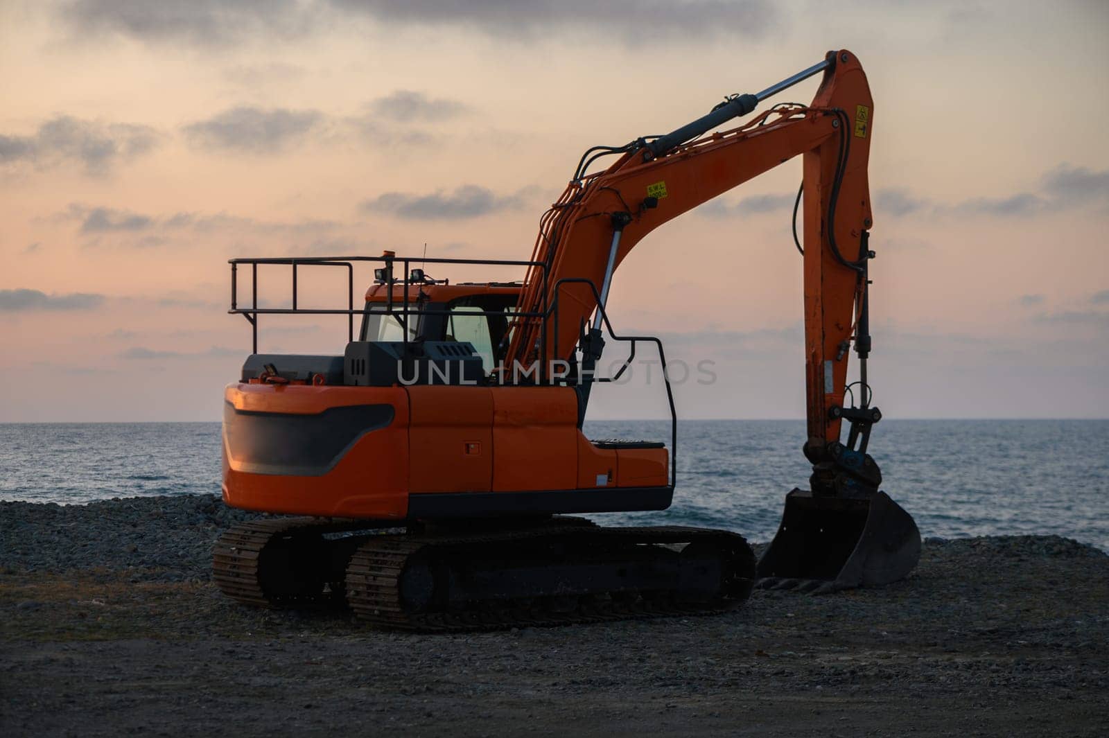 Heavy machinery stands ready at the water's edge, silhouetted against a vibrant sunset, indicating preparation for upcoming coastal construction tasks.