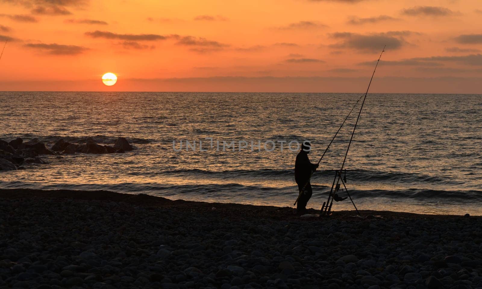 A silhouette of a dedicated fisherman stands on a rocky shore as the sun sets, painting the sky with warm hues. The rhythmic sound of waves completes this peaceful moment.