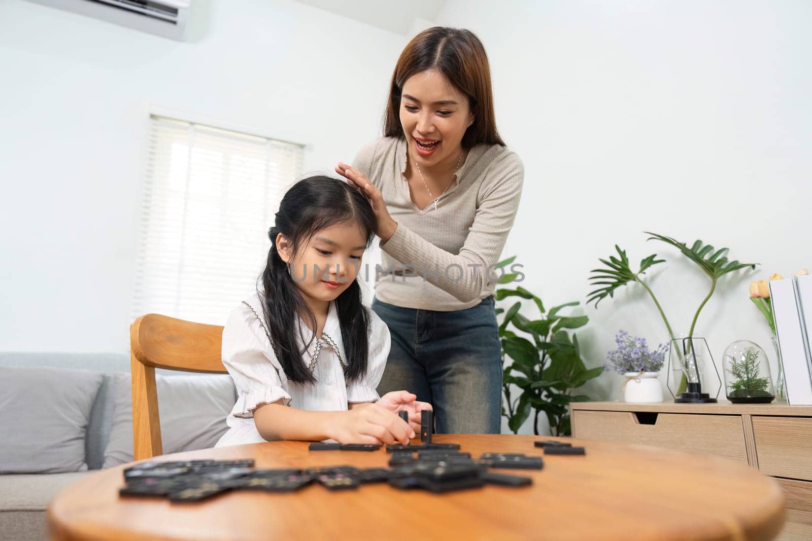 A mother gently encourages her daughter as they play a game of strategy with dominos, highlighting love, laughter, and developmental learning in a cozy home setting.