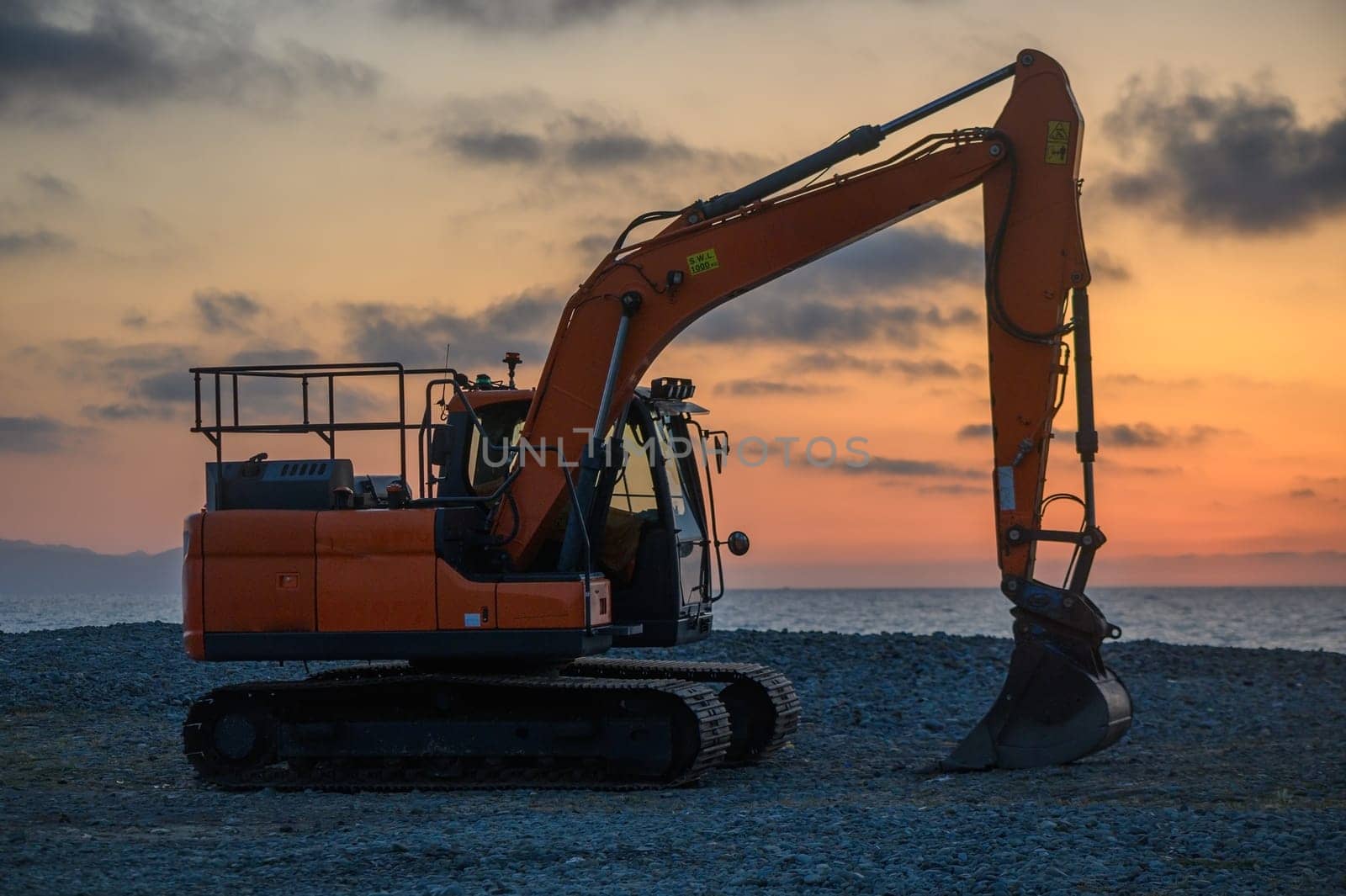 An orange excavator rests against a stunning sunset, silhouetted against the horizon. This machine is poised to shape the beach, creating new terrain by the sea.