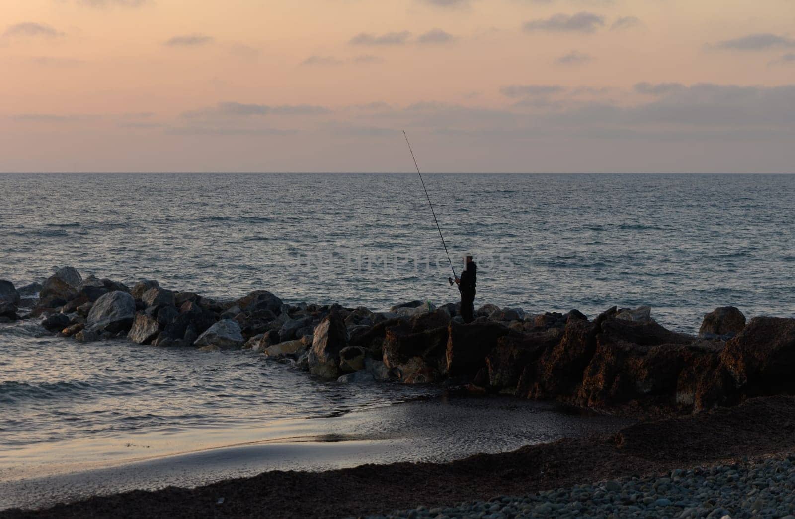 Silhouetted against a pastel sky, a lone fisherman stands on rocky shores, casting his line into the calm waters during the tranquil dusk hour.