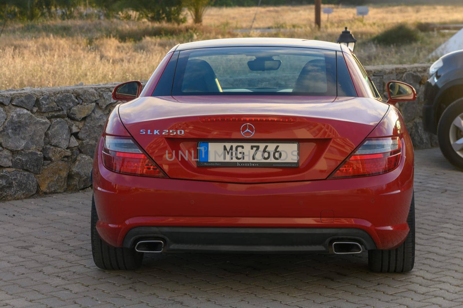 Gaziveren Cyprus -16.04.2025 A sleek red sports car stands parked near a rustic stone wall, with golden fields stretching in the background under a clear sky at sunset.