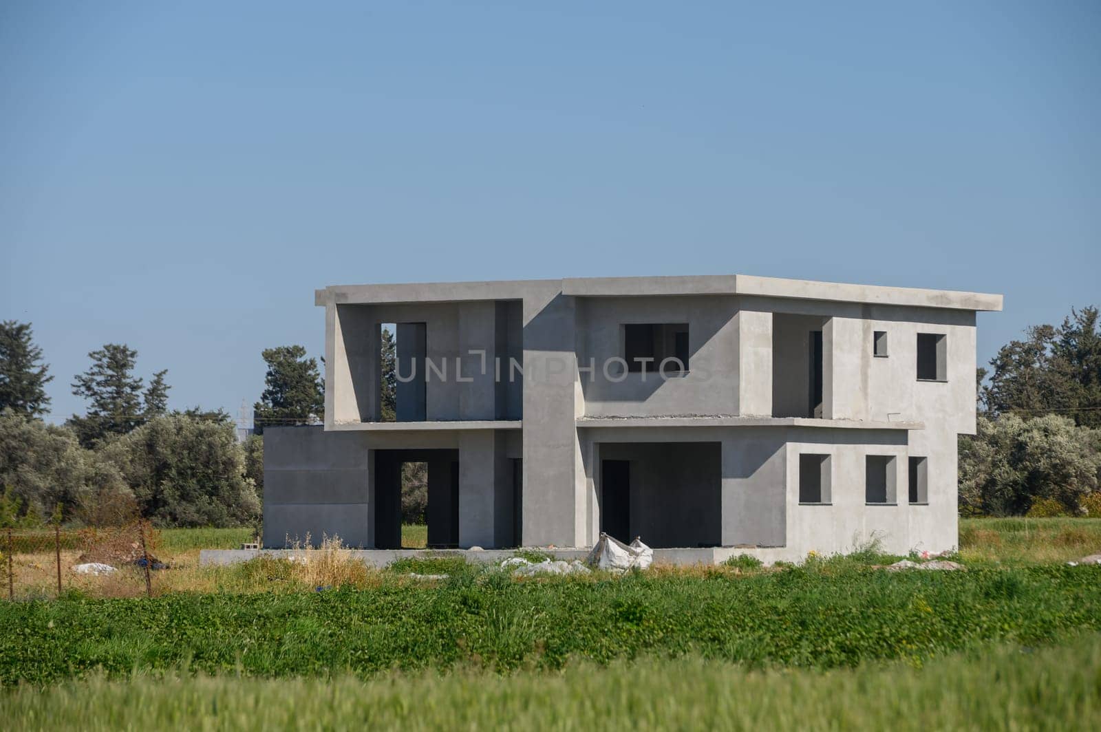 Amidst lush green fields, a minimalist concrete house stands tall, showcasing its modern architecture. The structure reflects contemporary design principles on a sunny day.