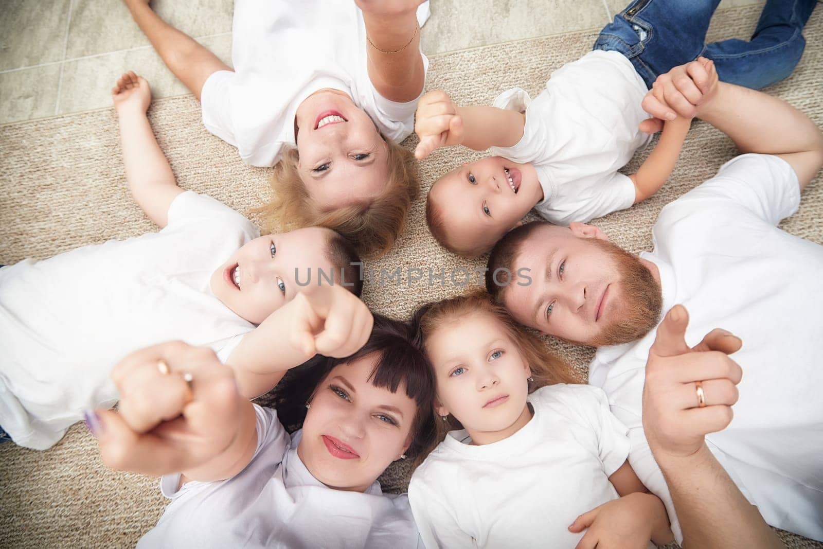 Six family members are lying on a rug, smiling and pointing at the camera, enjoying a fun moment together in a warm indoor setting.