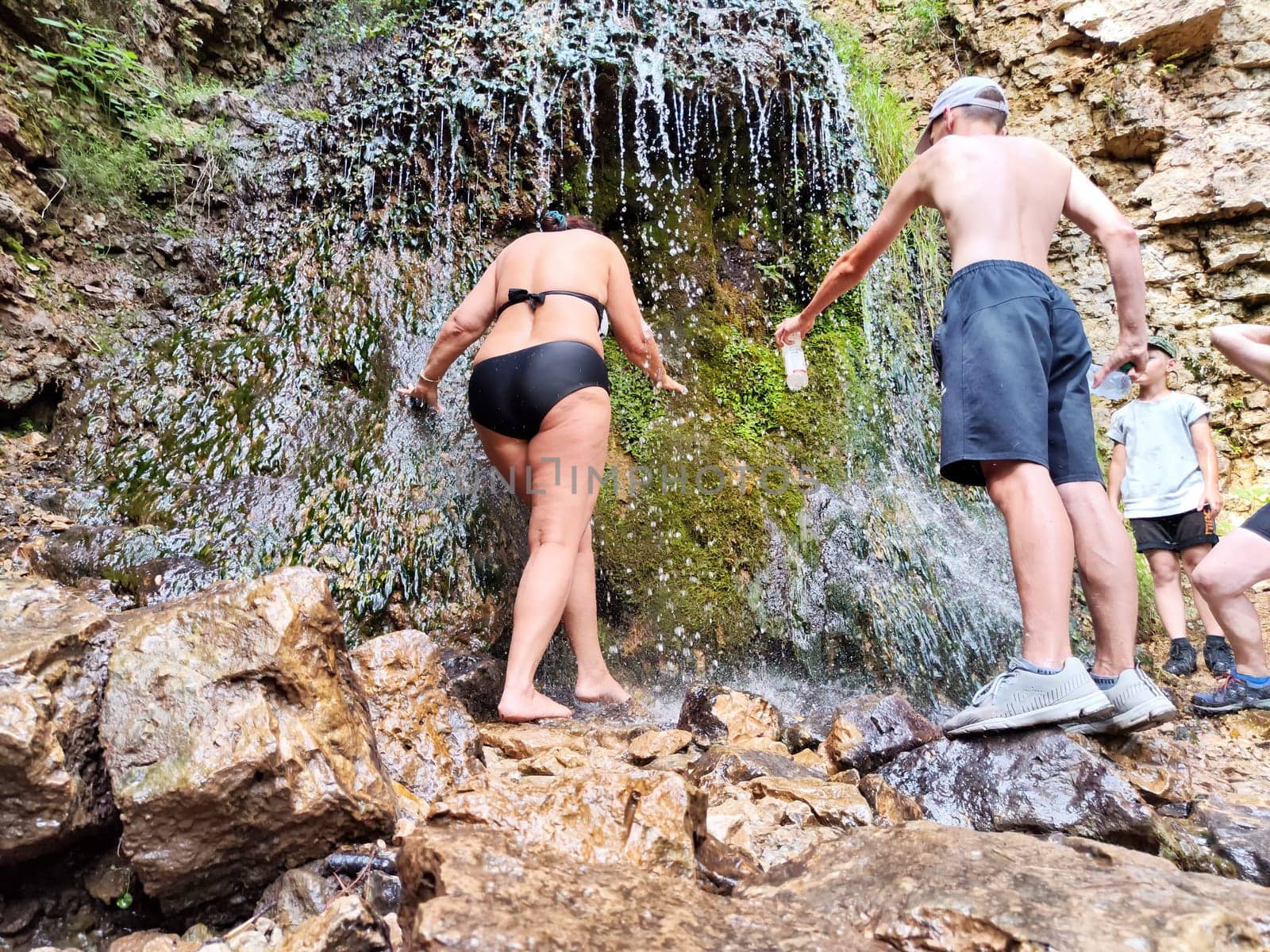 Friends enjoy a cooling experience under a waterfall in a rocky setting during a summer hike, surrounded by nature's beauty.