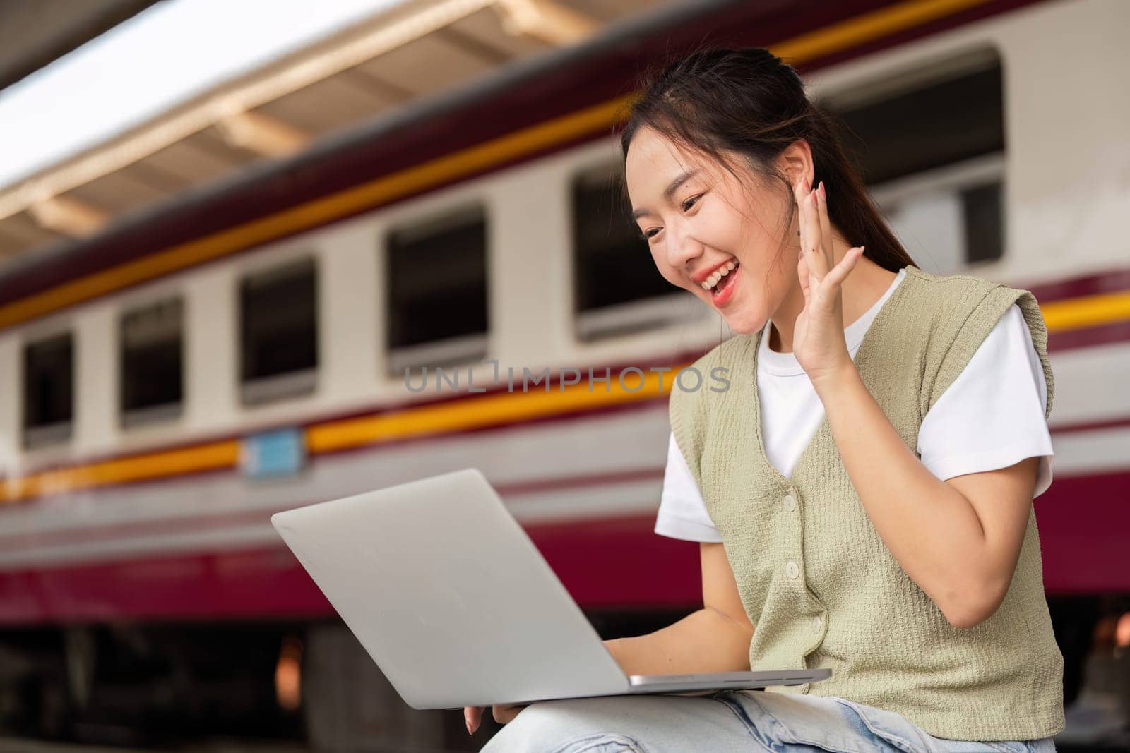 A joyful traveler sits at the train station, engaging in a video call on her laptop, illustrating the blend of travel and remote work in modern life.