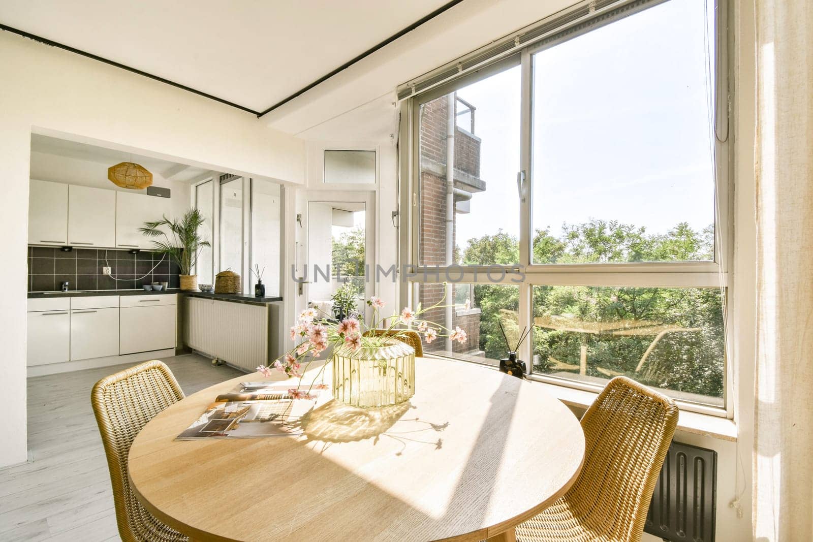 A sunlit dining area featuring a wooden table surrounded by woven chairs, with large windows offering views of greenery, perfect for a cozy dining experience.