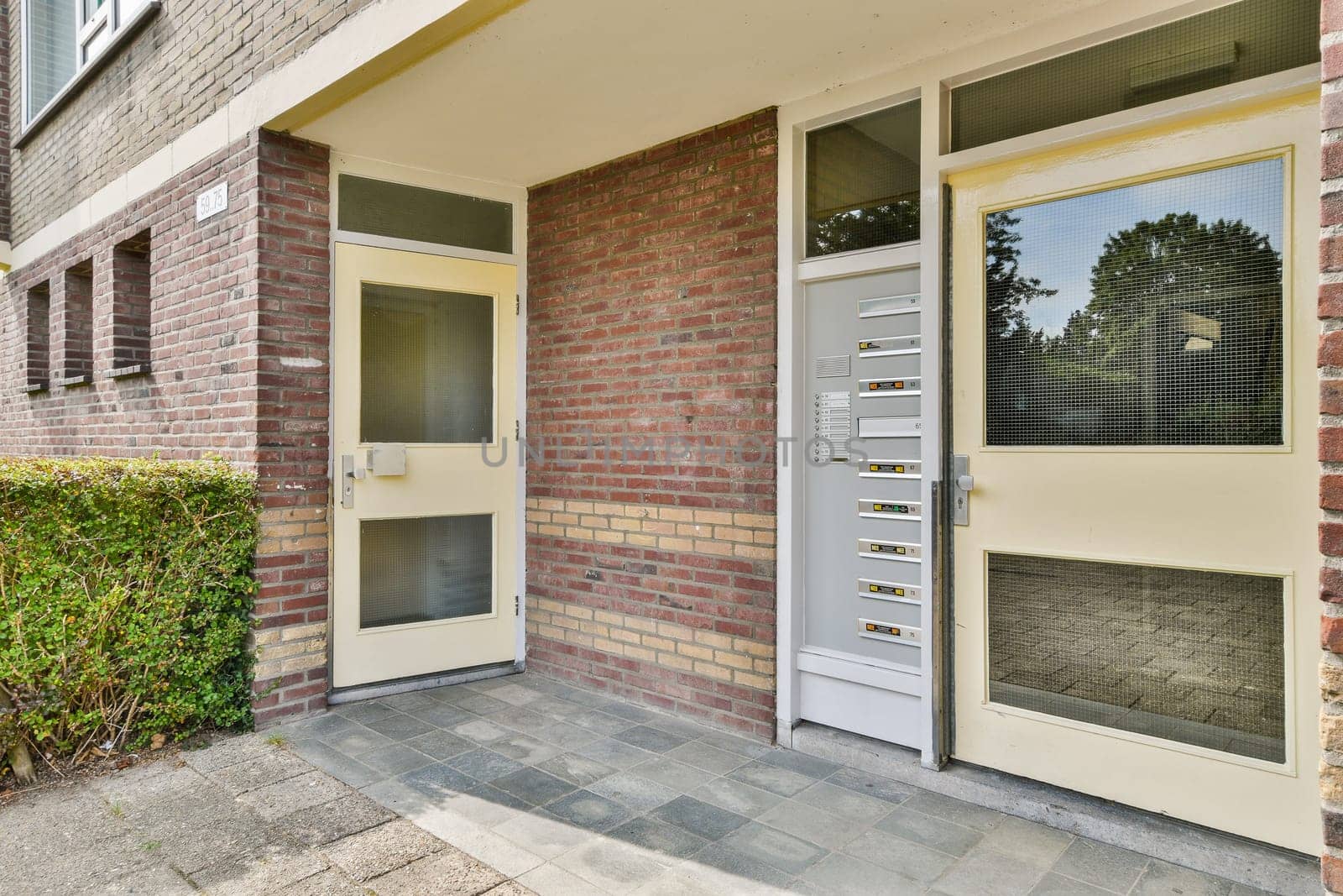 A spacious entrance area featuring contemporary doors and a brick facade, surrounded by greenery, indicating a welcoming residential environment.