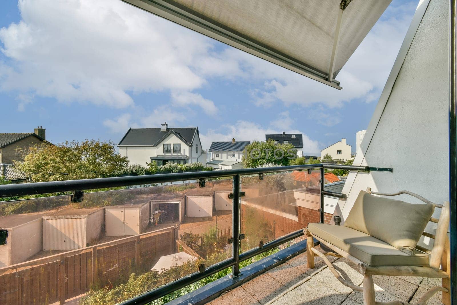 A serene balcony scene featuring a stylish chair and a view of residential houses surrounded by lush greenery and blue skies.