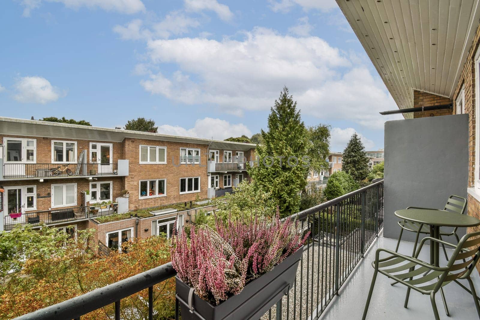 A scenic view from a balcony showcasing modern apartment buildings framed by greenery and vibrant flowers. The sunny sky adds to the inviting atmosphere.