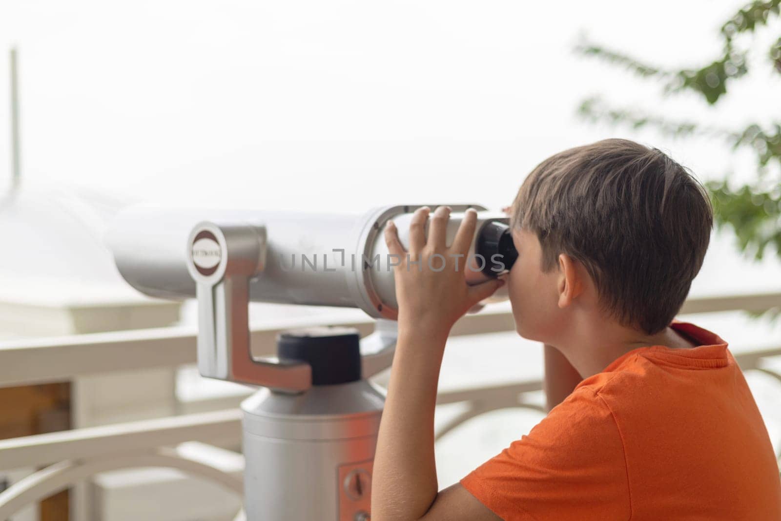Child in orange shirt using a mounted public telescope on an outdoor observation deck with railing and tree in the background
