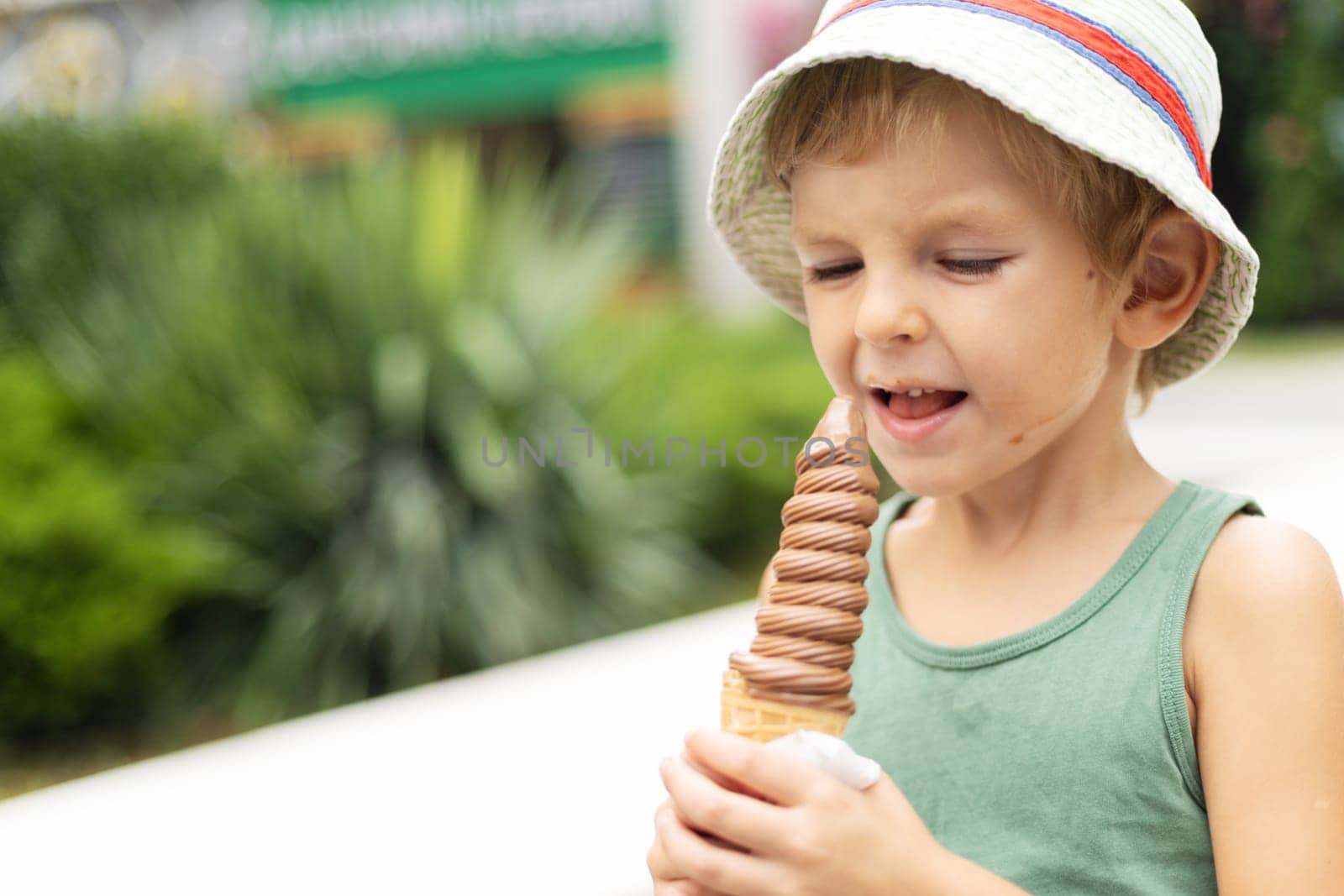 Young boy in green shirt and striped hat holding and licking chocolate ice cream cone on a sunny day