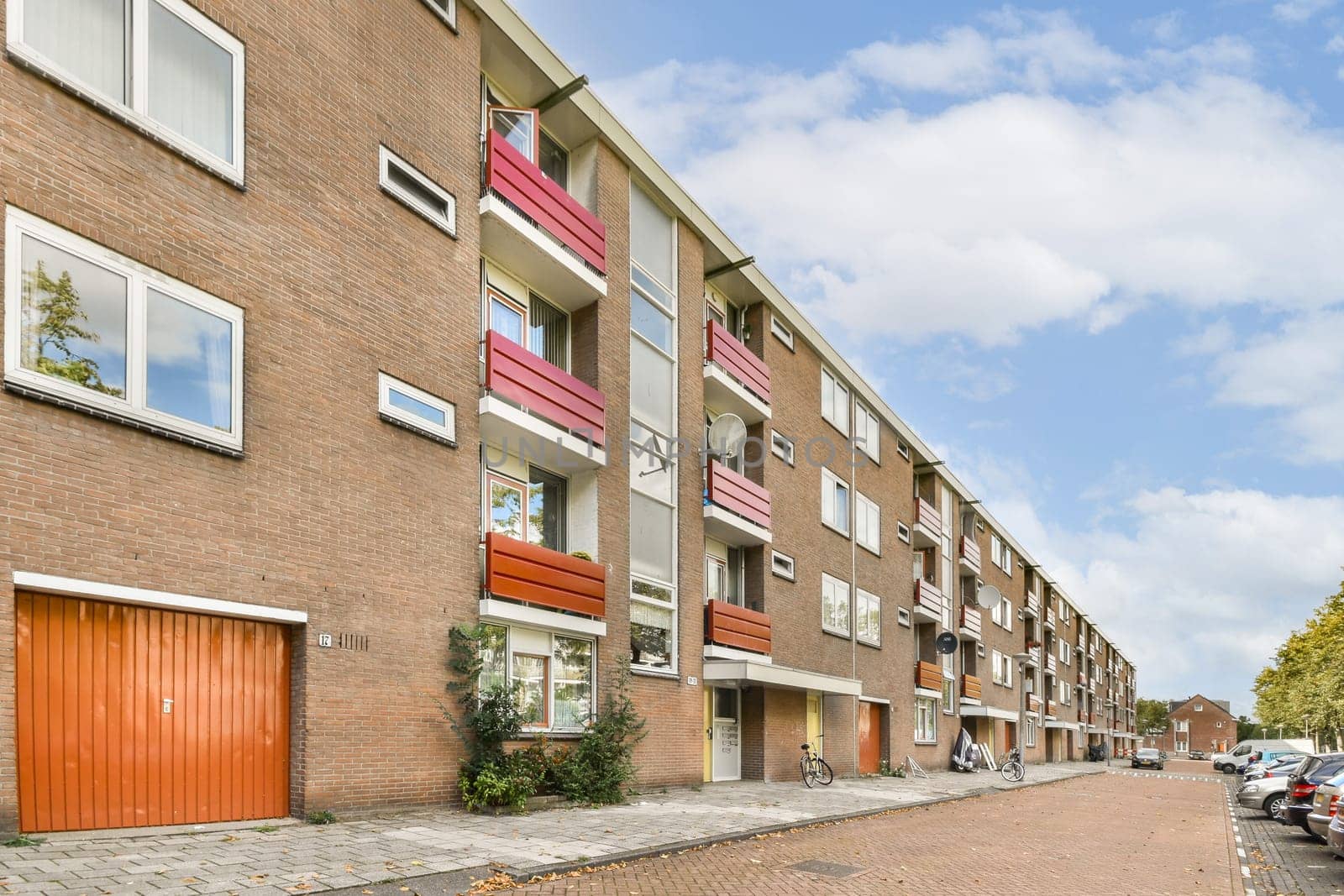 A modern residential building featuring brick architecture and vibrant balcony designs, located on a peaceful street lined with trees and parked bicycles.