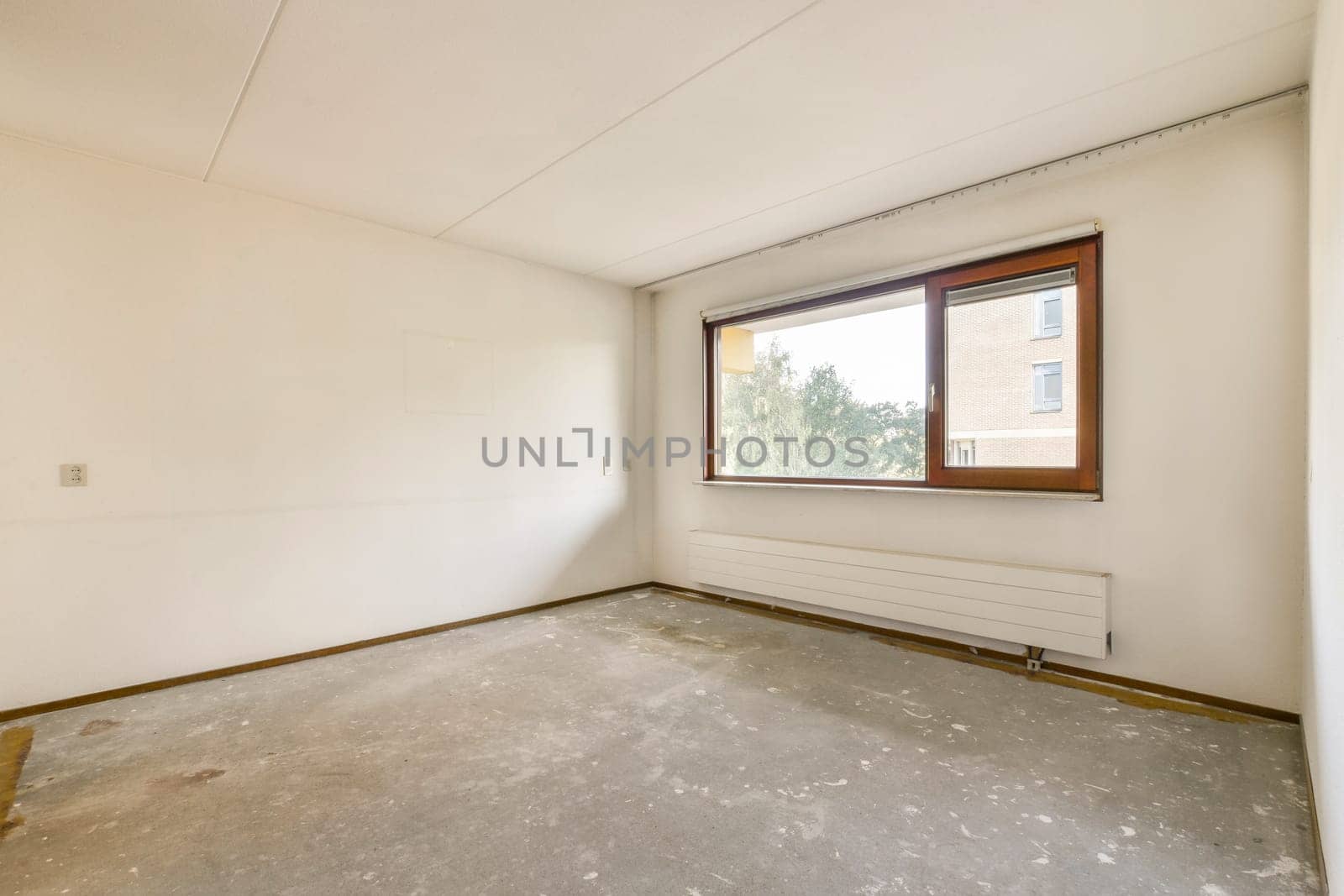 A spacious, empty room featuring a single window that lets in natural light, highlighting the unfinished walls and floor, creating a calm atmosphere for potential design.