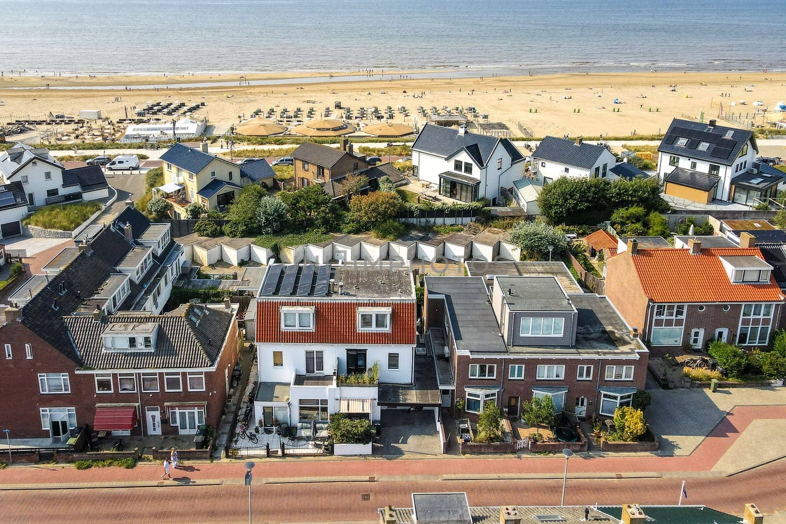 Aerial view showcasing coastal houses lined along a sandy beach, with umbrellas and sunbathers in the distance. Emphasis on architecture and natural beauty.