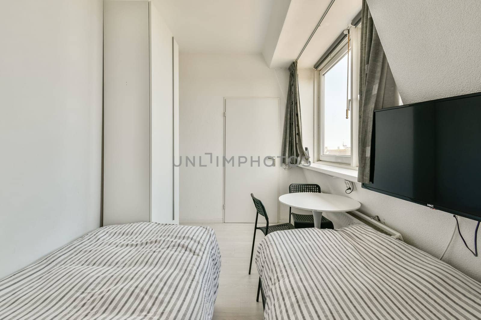 A bright and minimalist bedroom featuring two single beds, a table, and a television with large windows providing ample natural light.