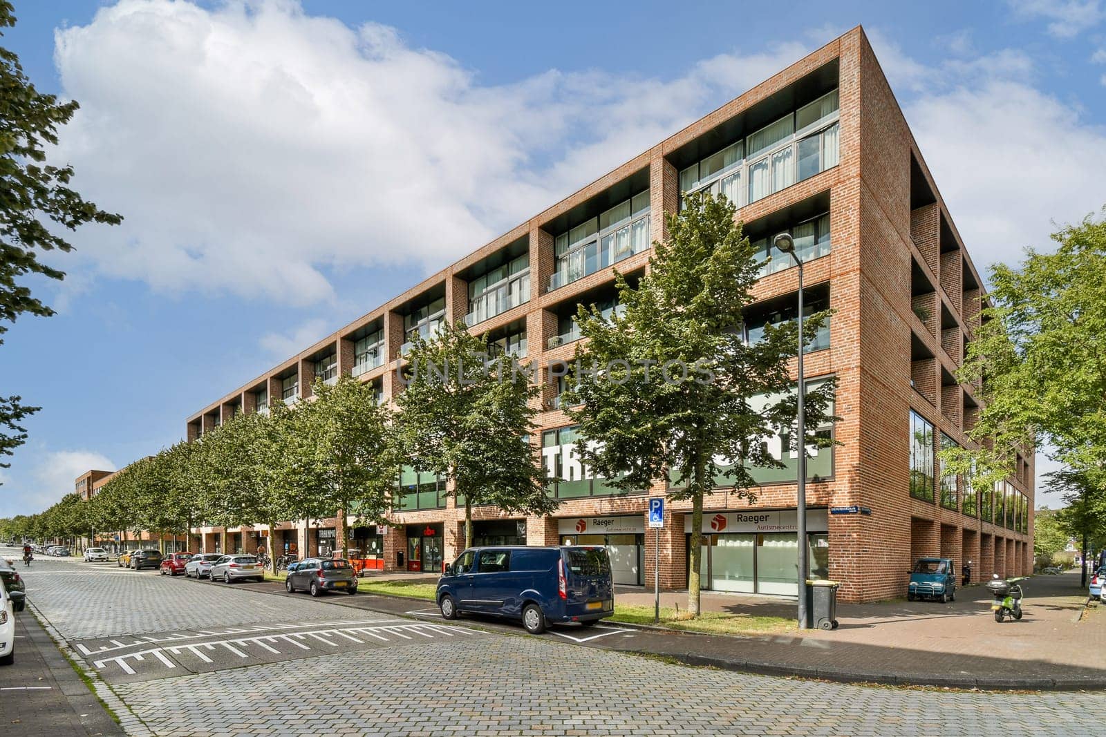 A contemporary building features large glass windows surrounded by lush greenery and trees, set on a bustling urban street with parked vehicles.