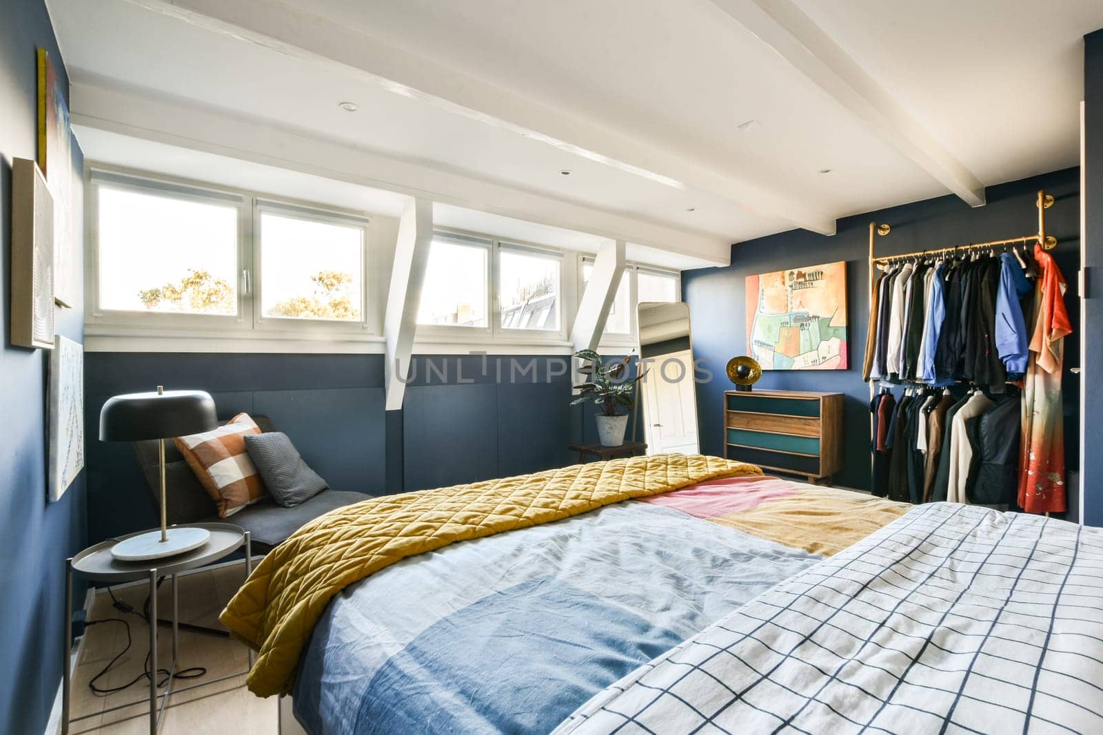 A contemporary bedroom showcasing a mix of textures and colors, featuring a cozy bed, stylish clothing rack, and beautiful natural light filtering through large windows.