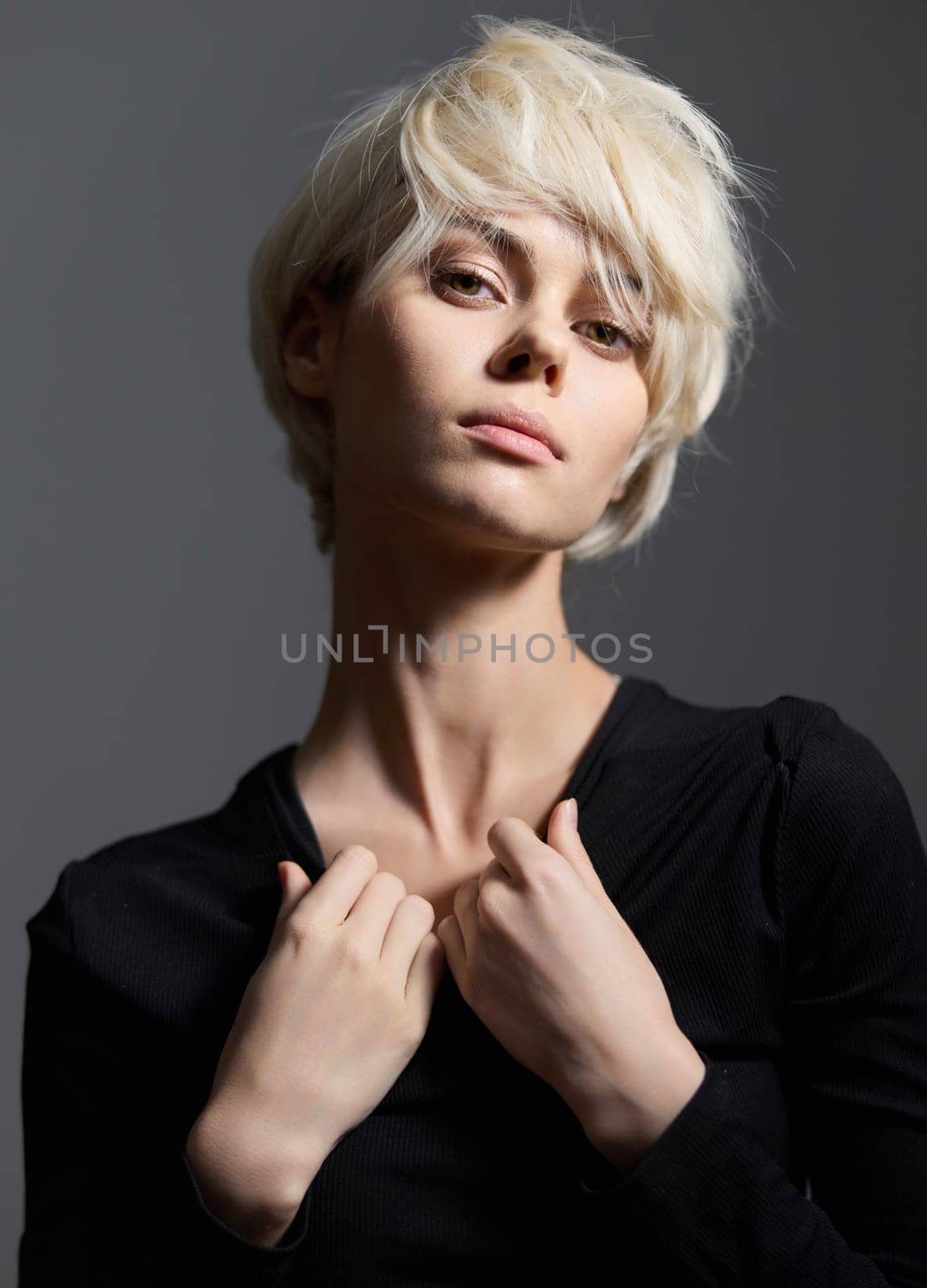 Elegant female model with short platinum blonde hair, wearing a fitted black top. Standing against a dark gray background, she portrays strength and self-assurance through her intense gaze and postur