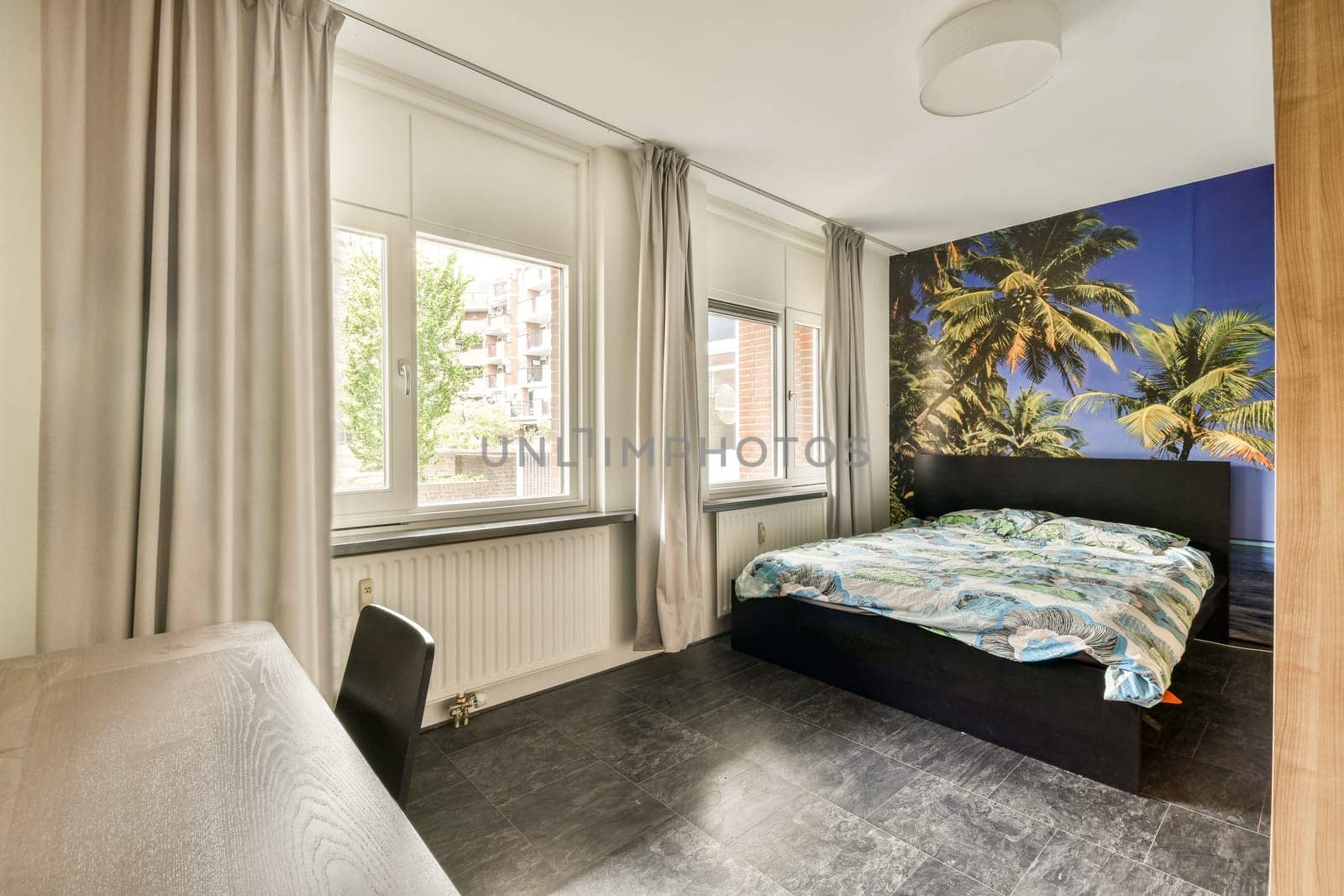 A stylish bedroom featuring a large bed adorned with a colorful duvet, complemented by a tropical mural and natural light flowing through the windows.