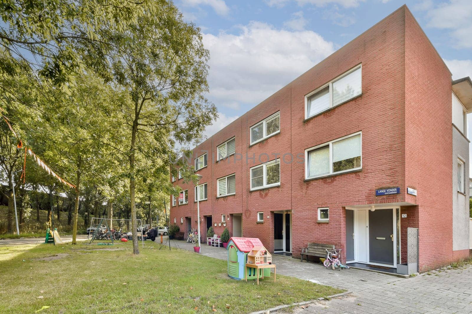 A contemporary residential building showcasing a red brick facade, surrounded by greenery and playground equipment, ideal for family living.