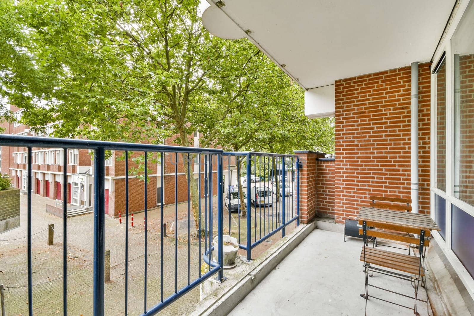 A serene balcony view featuring a wooden table and chairs, surrounded by leafy trees and a peaceful neighborhood street with parked cars.