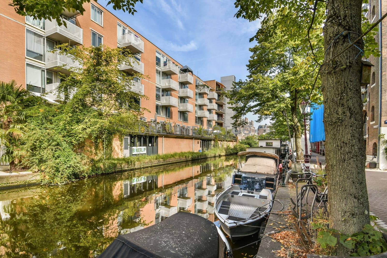 A picturesque canal scene showcasing modern buildings alongside lush greenery, with boats lined along the waterway, reflecting the architecture beautifully.