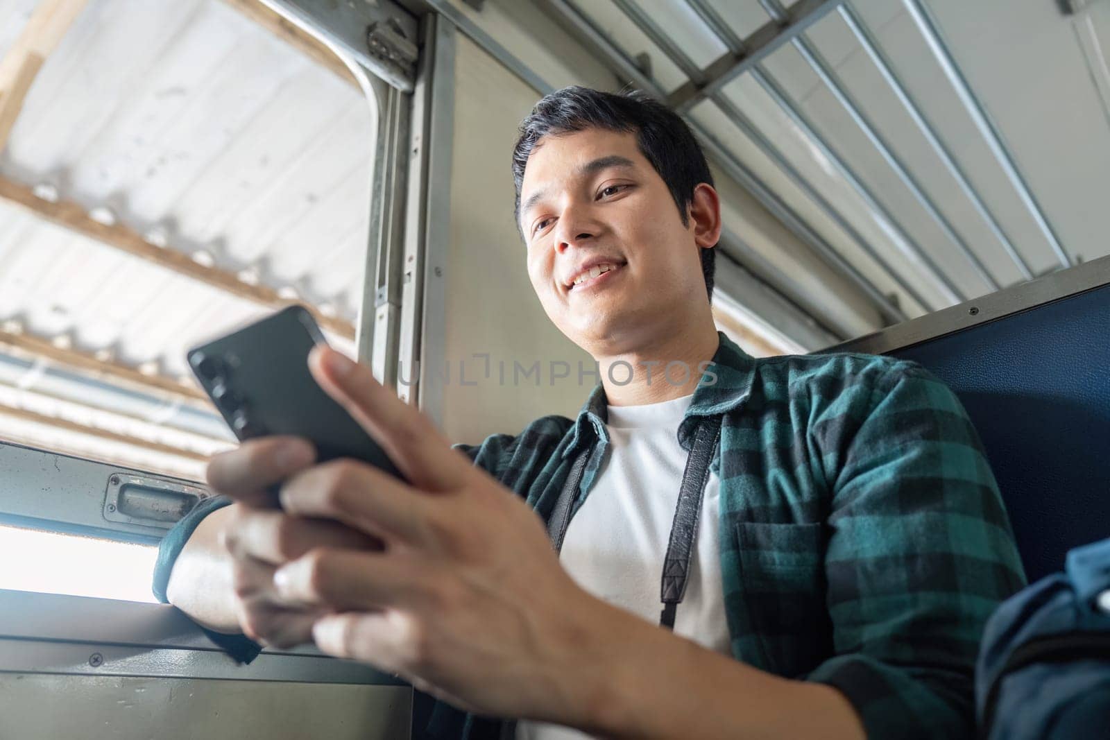 Relaxing on the train, a young traveler smiles while using his smartphone to connect with friends and share his journey experiences.