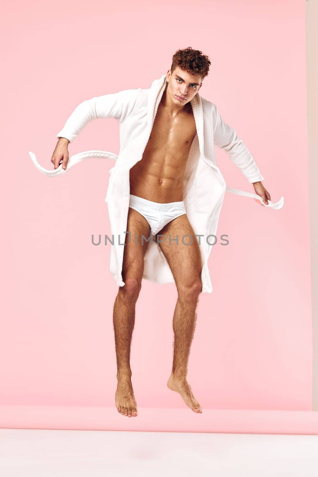 Energetic young man showcasing a stylish white robe and underwear, jumping playfully against a pink backdrop. Concept of freedom and contemporary men's fashion.