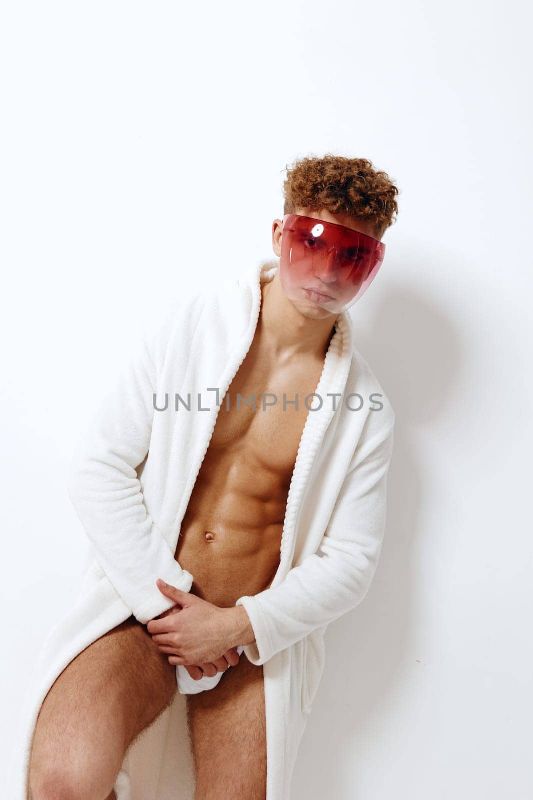 Confident young male model wearing a fluffy white robe and vibrant sunglasses. The neutral backdrop emphasizes his bold fashion choice and contemporary style.
