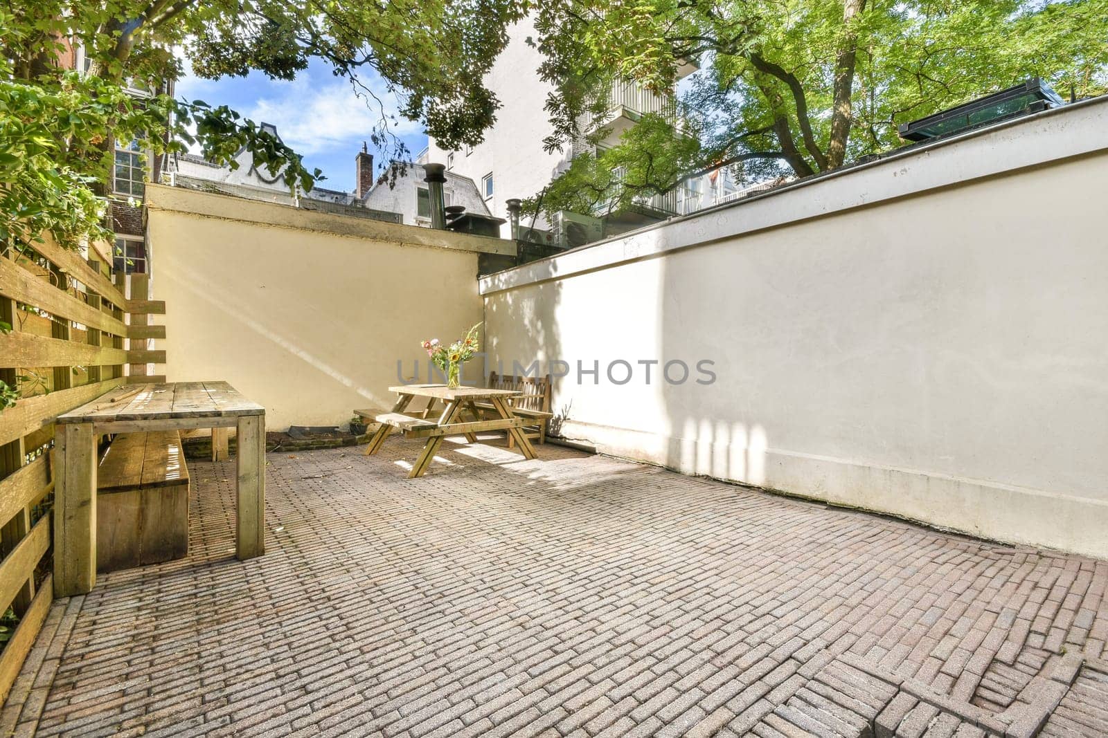 A serene outdoor patio featuring light brick flooring, wooden furniture, and lush greenery, perfect for relaxation and social gatherings.