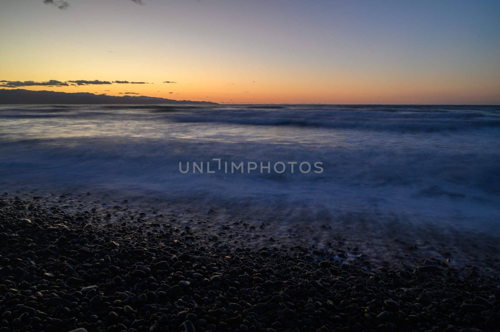 Waves crash gently against a rocky shoreline as the sun sets, painting the sky with hues of orange and purple. A serene atmosphere envelops the coastal landscape, inviting reflection.