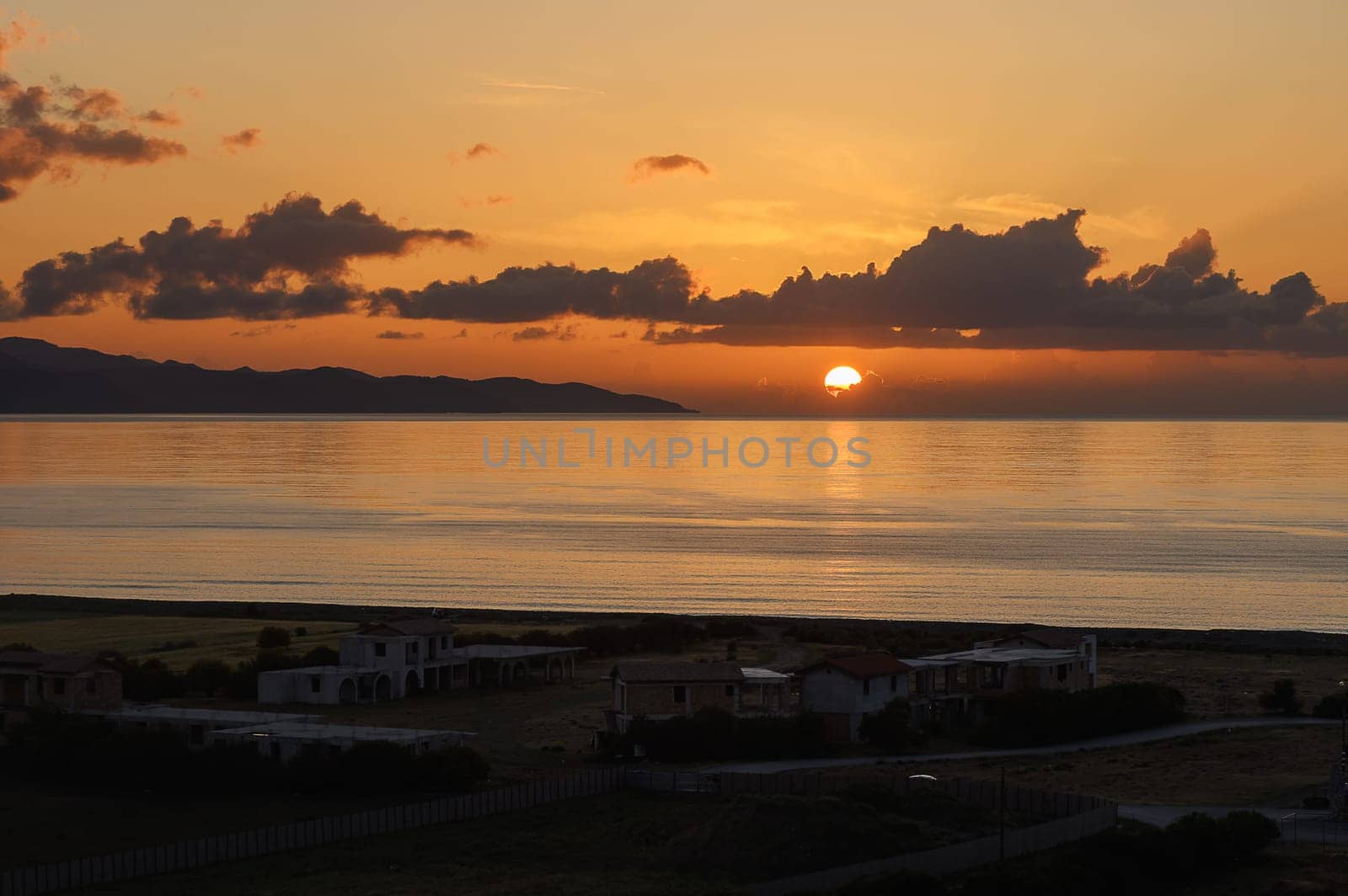 As the sun descends towards the horizon, it paints the sky with warm oranges and pinks, reflecting upon calm waters. A quiet village rests peacefully, framed by gentle waves and distant hills.