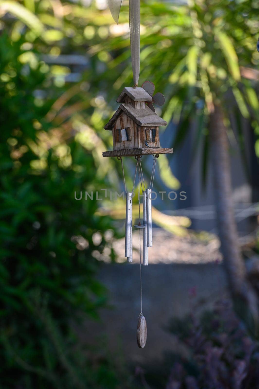 A beautifully crafted wooden wind chime shaped like a house softly sways in the afternoon breeze, surrounded by vibrant tropical plants.
