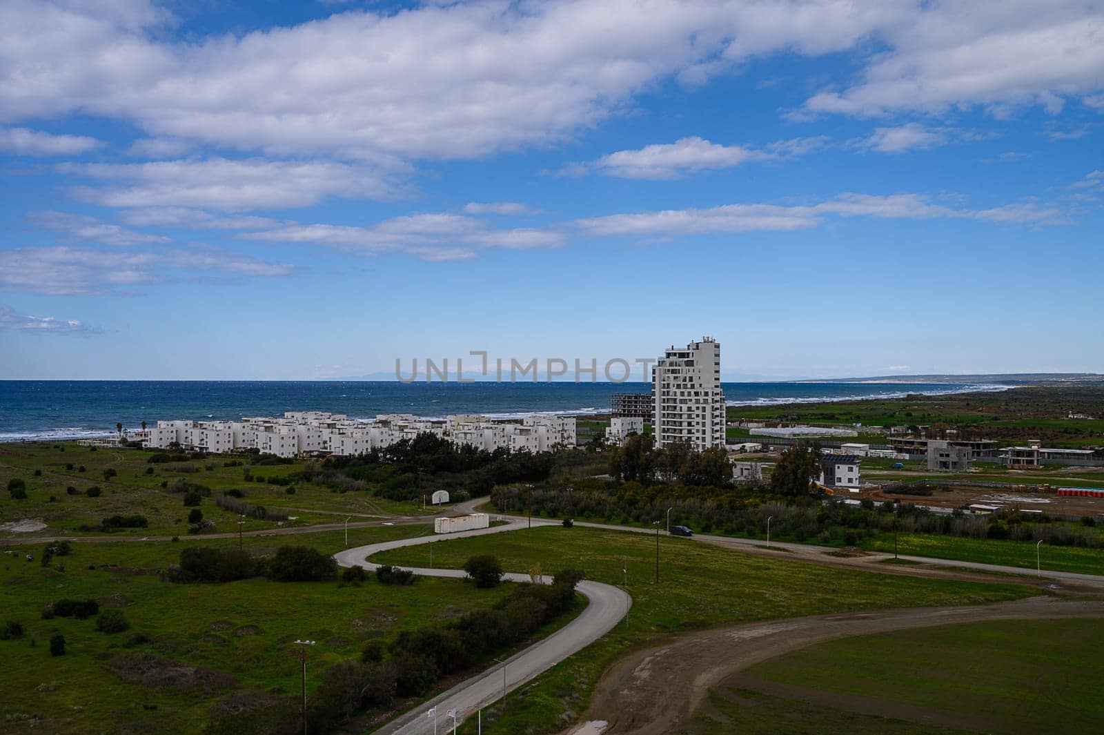 Beneath a brilliant blue sky, sleek buildings edge the coastline, with lush greenery and winding roads leading to the sparkling ocean. A serene harmony of nature and modernity blooms.