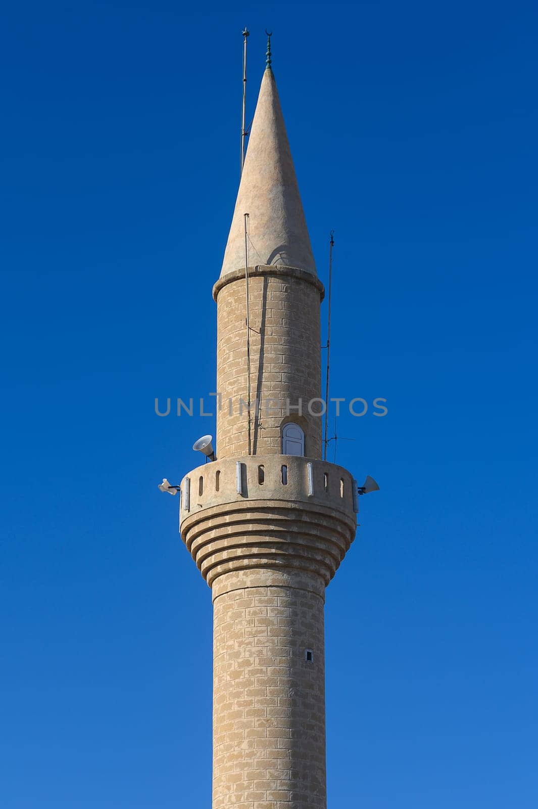 A striking minaret rises gracefully, showcasing intricate stonework and a tall spire. Nestled in a lively town, it stands as a testament to rich cultural heritage under a bright blue sky.