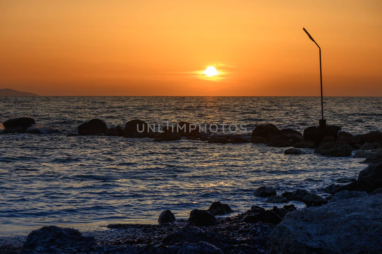 Vibrant hues of orange and gold fill the sky as the sun dips below the horizon. Gentle waves lap against the rocks while a solitary post stands quietly by the shore.