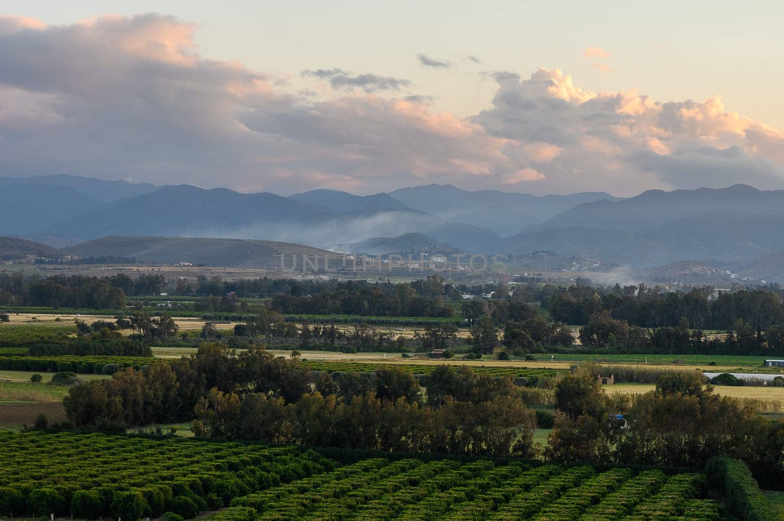A breathtaking view captures the tranquility of lush green fields under a pastel sky as the sun sets behind distant mountains, casting a warm glow over the peaceful countryside.