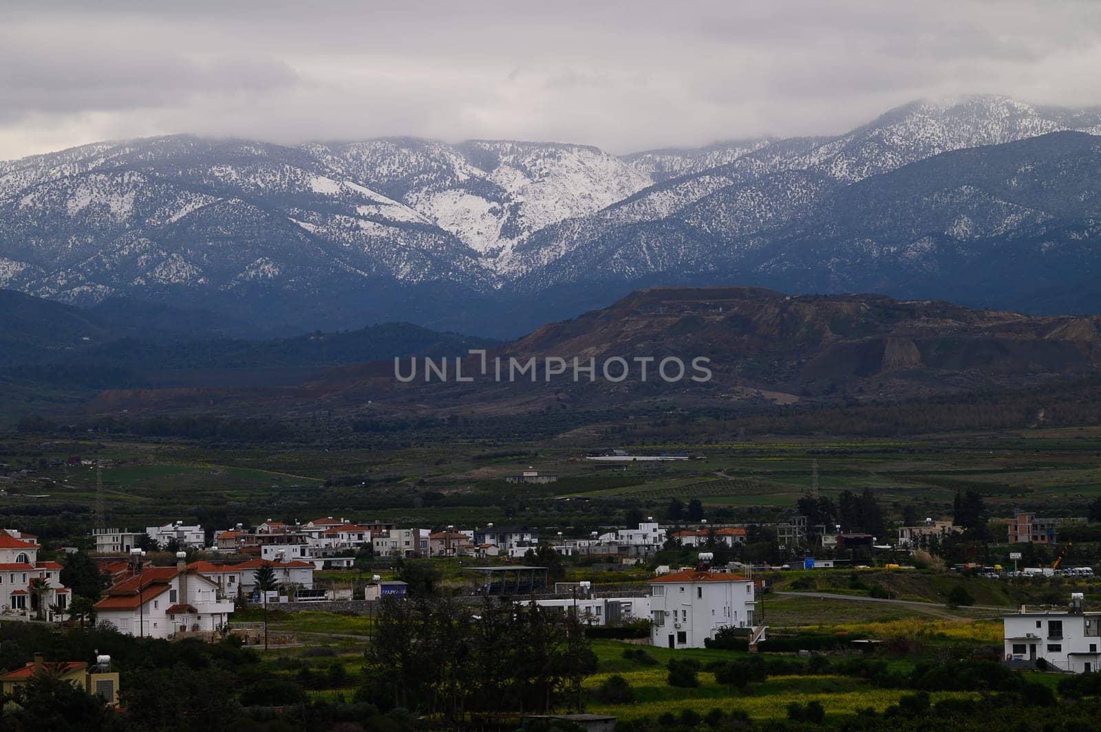 Snow-covered peaks rise majestically above a tranquil village, dotted with white and red-roofed houses amid vibrant green fields on a cloudy day.