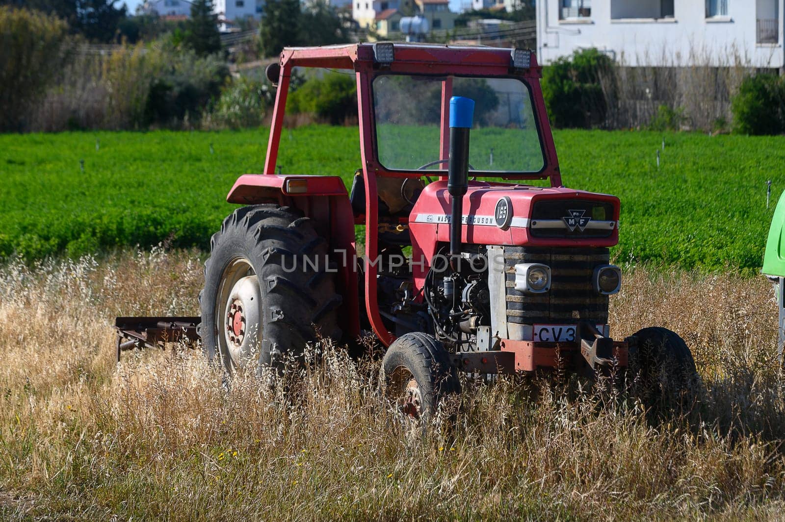Gaziveren Cyprus -10.04.2025 An abandoned red tractor sits in a lush green field surrounded by golden grass. The bright afternoon sun casts shadows, revealing the beauty of rural life and nature's harmony.