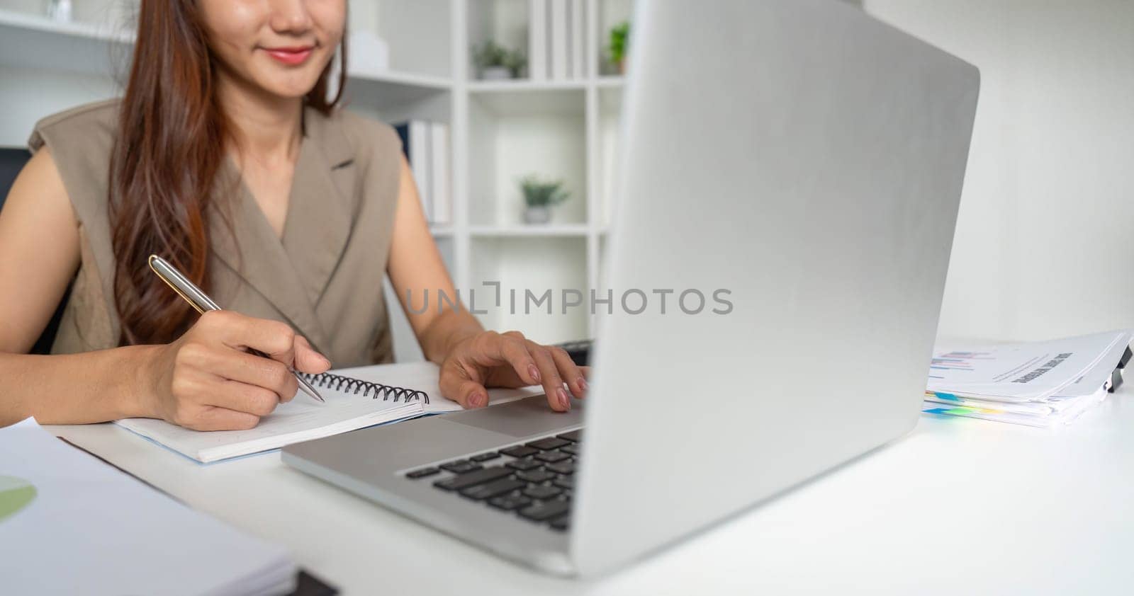 A woman confidently jots down notes as she engages in her work on a laptop, showcasing an organized and productive work atmosphere.