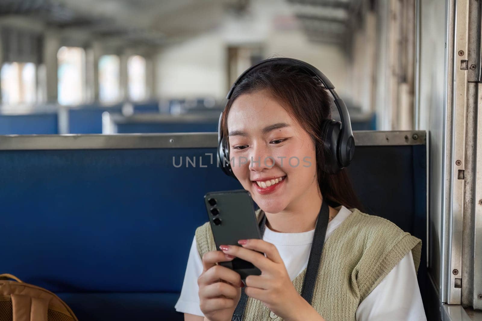 A cheerful traveler wearing headphones smiles while using her smartphone to listen to music or watch videos during her train journey, reflecting moments of leisure and adventure.