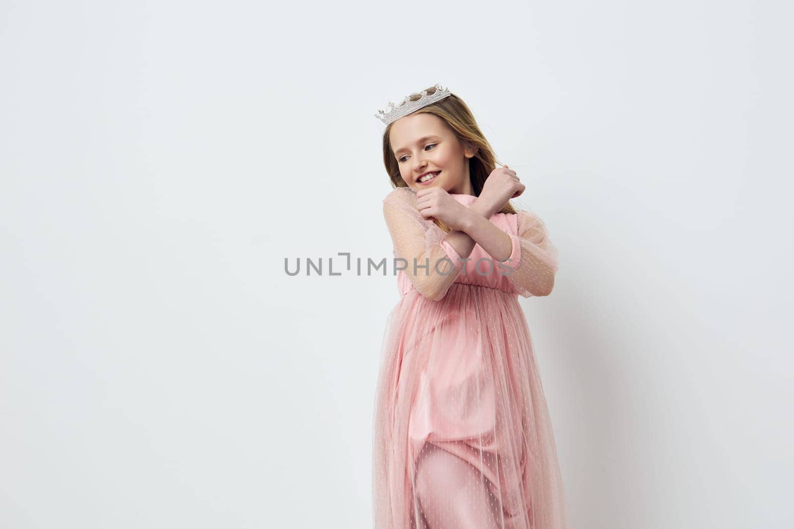 This image features a cheerful young girl dressed in a beautiful pink tulle dress and tiara, playfully crossing her arms against a clean white backdrop The soft pastel colors evoke a sense of innocence and joy, ideal for children s fashion or celebratory themes
