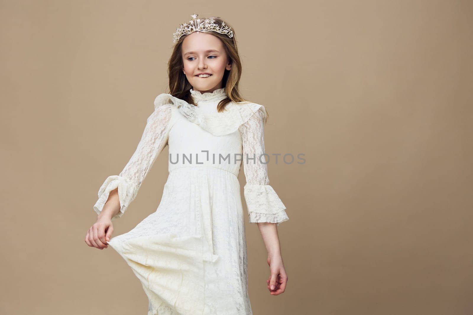 A joyful young girl in a white lace dress and tiara, embodying innocence and playfulness against a neutral backdrop, perfect for themes of childhood elegance and celebration